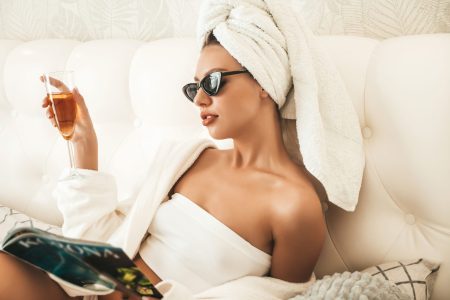Young beautiful smiling female in white lingerie and towel on head. Sexy carefree woman lies on bed in posh apartment or hotel room. Hot model after shower reading magazine and drinking champagne