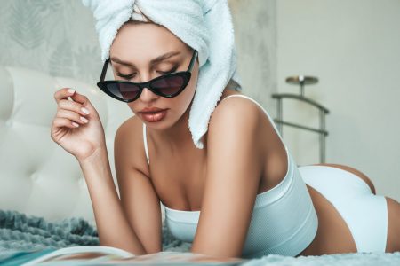Young beautiful smiling female in white lingerie and towel on head. Sexy carefree woman lies on bed in posh apartment or hotel room. Hot model after shower reading magazine. Enjoying her morning